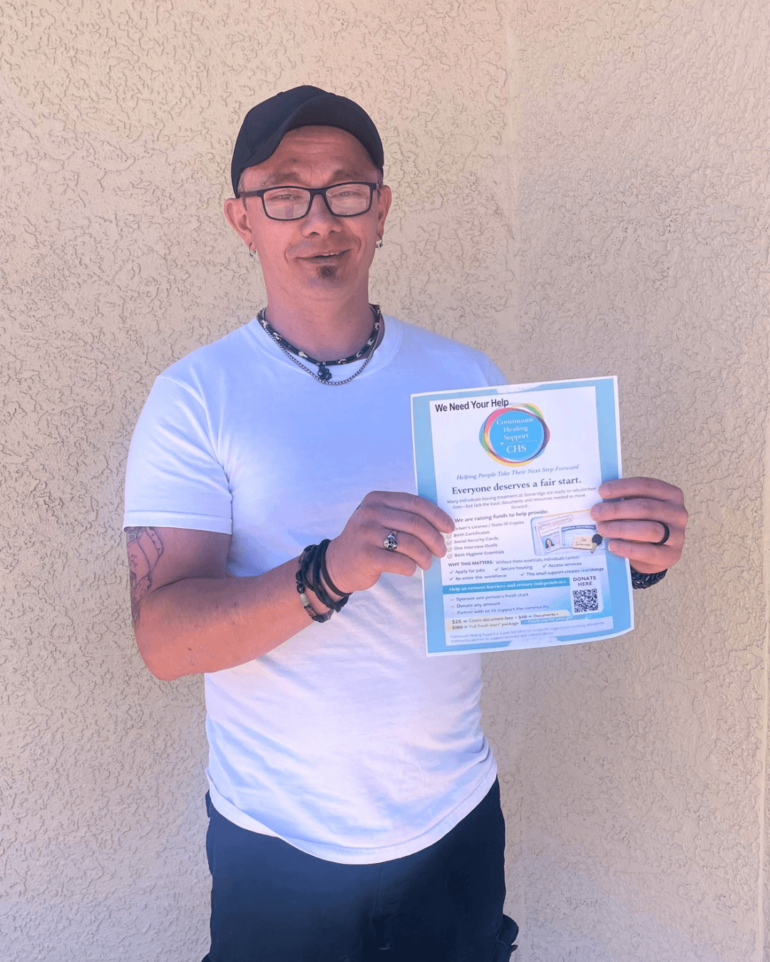 Smiling man in a white t-shirt and black cap holding a Continuum Healing Support flyer.